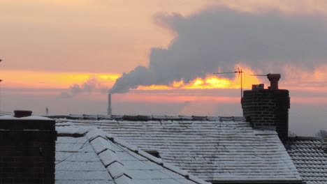Smoking-industry-chimney-above-frosty-wintery-home-rooftops-glowing-sunrise-orange-sky