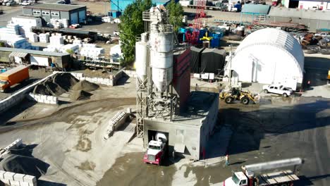 Aerial-pan-left-shot-of-Silos-on-a-Industrial-Complex-with-Semi-Trucks,-Humans-working-and-heavy-equipment-moving