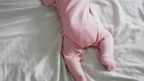 Young-baby-with-pink-outfit-trying-to-crawl-on-white-bed,-top-down-view