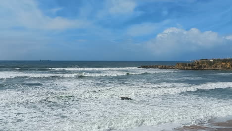 Rough-sea-creating-several-waves-of-white-foam,-some-brave-surfers-and-the-blue-sky-with-lots-of-clouds