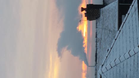 VERTICAL-clip:-Industry-smoking-chimney-above-frosty-winter-residence-rooftops-during-orange-glowing-sunrise