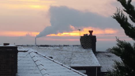 Smoking-energy-industry-chimney-above-frosty-winter-home-rooftops-glowing-sunrise-orange-sky