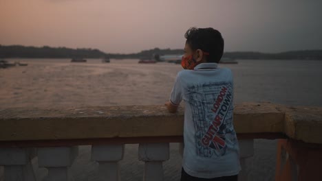Eine-Nahaufnahme-Eines-Begeisterten-Kleinen-Jungen,-Der-Auf-Verschiedene-Boote-Hinweist,-Die-Hinter-Einer-Betonbalustrade-Stehen,-Die-Den-Schönen-Mandovi-fluss-Bei-Sonnenuntergang-überblickt,-Goa,-Indien