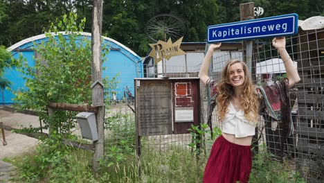 Beautiful-woman-Eco-Conscious-Hippie-Holding-Onto-And-Looking-At-anticapitalist-street-sign-Post-Saying-Kapitalisme-weg,-away-with-capitalism,-postcapitalism,-back-to-basics-in-nature,-off-grid-living
