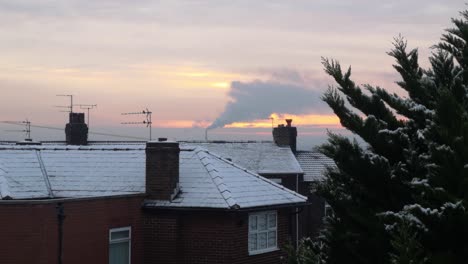 Smoking-industry-chimney-above-snowy-winter-home-rooftops-glowing-sunrise-orange-sky