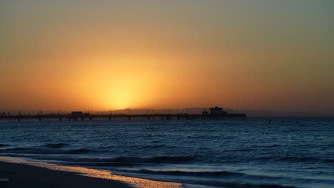 Silhouette-of-a-pier-during-a-golden-sunrise-with-the-waves-gently-washing-in-over-a-calm-beach---time-lapse