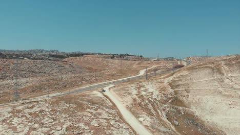 Drohne-Geschossen-In-Der-Judäischen-Wüste,-Braune-Hügel,-Sandweg,-Autos,-Die-Auf-Der-Straße-Fahren,-Strommasten-Und-Kleinstädte-Weit-Entfernt,-Israel