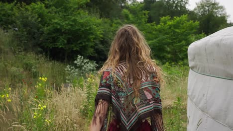 Back-Of-Female-Eco-Conscious-Hippie-Walking-Through-Wild-Gardens-Next-To-Yurt-in-ecovillage