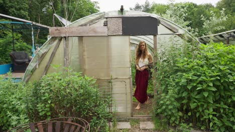 Caucasian-Hipster-girl-in-her-20th-walk-out-of-handmade-village-greenhouse-with-may-green-plants-around---slow-motion
