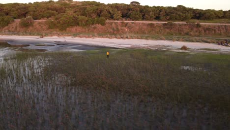 Aerial-view-of-man-with-taking-photos-with-mobile-phone-between-wet-grass-at-lagoon-shore-during-sunset-time---Black-Lagoon,Uruguay