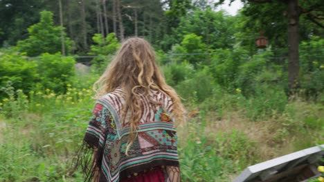 Free-hippie-young-person-walking-back-to-camera-through-wild-flowers-field-in-nature,-organic-sustainable-agriculture-ecovillage-tinyhouse-lifestyle