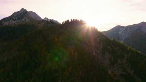 Vista-Aérea-Que-Se-Eleva-Sobre-El-Pico-De-La-Montaña-Del-Bosque-Alpino-Del-Tirol-Austríaco-Para-Revelar-El-Sol