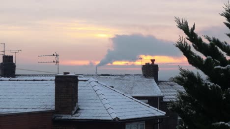Smoking-industry-chimney-above-frosty-white-winter-home-rooftops-glowing-sunrise-orange-sky