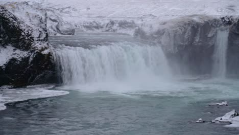 Panning-shot-of-Godafoss-waterfall-crashing-down-during-frosty-cold-day-in-Winter,-Iceland---Icebergs-floating-in-water