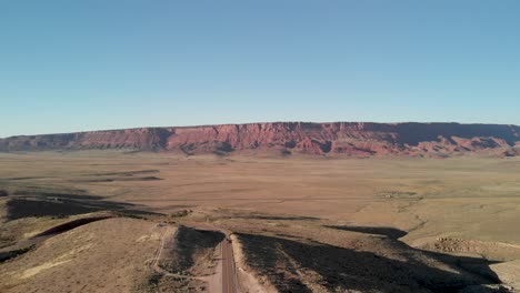 Aerial-shot-of-an-empty-road-going-into-the-distance-to-red-mountain-rocks-in-canyon-landscape-in-the-dried-desert