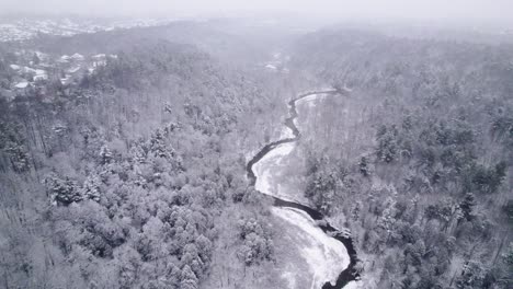 Winding-River-cuts-through-a-Snow-Covered-Valley-during-a-Blizzard-with-Fresh-snow,-White,-Aerial-Drone,-Snowing-in-Winter-Canada