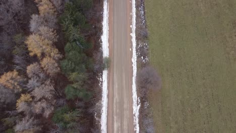 Aerial-Shot-Of-A-Dirt-Road-Lined-With-Snow,-Forest-And-Farm-Land