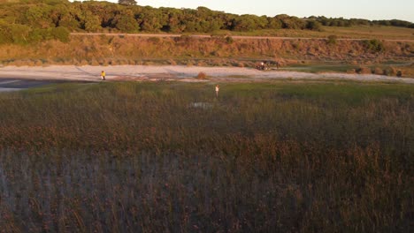 Aerial-tracking-shot-of-redhead-woman-walking-on-grassy-shore-of-nature-Lagoon-at-sunset---Uruguay,South-America