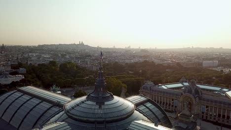 Drone-tour-of-Paris-city-from-above,-with-French-flag-waving-on-top-of-Grand-Palais-and-Petit-Palais-at-the-dawn,-highlighting-silhouette-view-of-Sacre-Coeur-at-distance