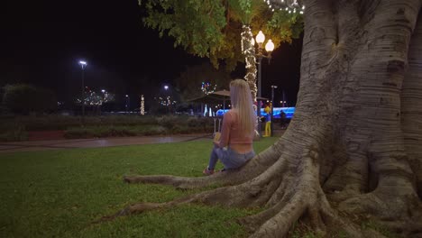 Behind-blond-woman-sitting-on-tree-root-in-park-at-night-eating-chestnuts,-Funchal