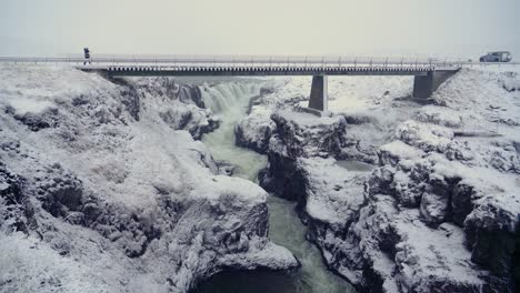 Man-Walking-Across-A-Bridge-In-Kolugljufur-Canyon-Iceland-In-Winter---wide-shot