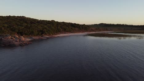Aerial-flight-over-tranquil-Black-Lagoon-with-sandy-beach-and-forest-trees-during-sunset-in-Uruguay---Wild-Birds-flying-at-sky