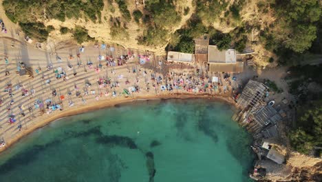 Cinematic-aerial-shot-of-tourists-relaxing-and-sunbathing-at-the-turquoise-seashore-of-Cala-Benirras-in-Spanish-island-of-Ibiza