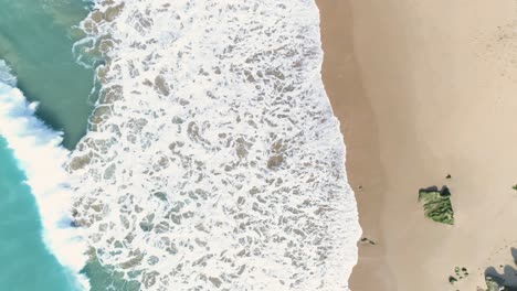 Aerial-view-of-powerful-turquoise-blue-sea-waves-breaking-on-the-sand-of-a-beautiful-white-sand-beach-with-surfers-in-the-water