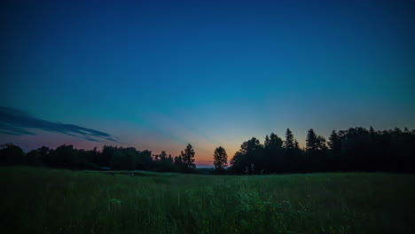 Schöner-Sonnenaufgang-Einer-Wiese-Mit-Wald-Im-Hintergrund-Unter-Einem-Blauen-Himmel-Mit-Gelben-Und-Orangefarbenen-Farben-In-Den-Vorbeiziehenden-Wolken