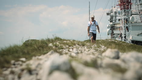 Hiker-with-a-cap-and-an-orange-backpack-walking-away-from-the-radio-tower-on-rocky-path-with-patches-of-grass