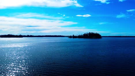 parallel-summer-aerial-over-loon-bird-habitat-lake-reflective-view-of-blue-sky-clouds-with-a-natural-island-in-the-center-surrounded-by-a-forest-park-with-sparkling-water-as-the-sun-sets-gradation