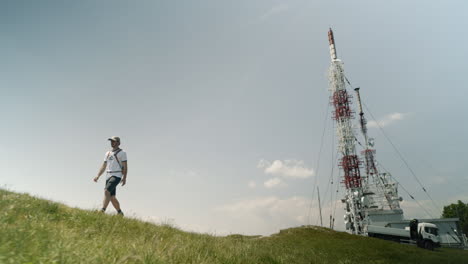 Hiker-with-a-cap-and-backpack-walking-away-from-a-radio-tower-on-mountain-Nanos-on-a-summer-day