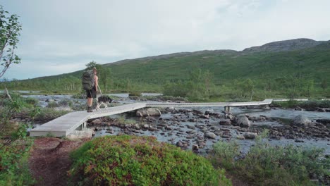 Caucasian-Guy-Hiking-With-A-Husky-Dog-Crossing-A-Rocky-River-On-Wooden-Footpath-At-Anderdalen-National-Park-In-Senja,-Norway