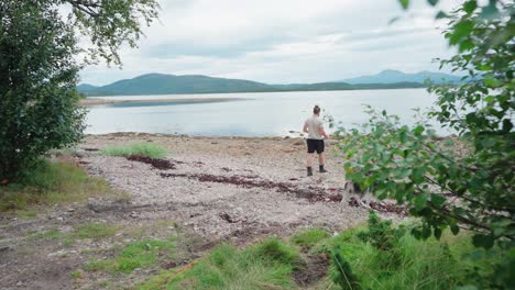 Un-Hombre-Caminando-Con-Su-Perro-Hacia-La-Orilla-Rocosa-Del-Río-En-El-Parque-Nacional-Anderdalen,-Isla-Senja,-Noruega