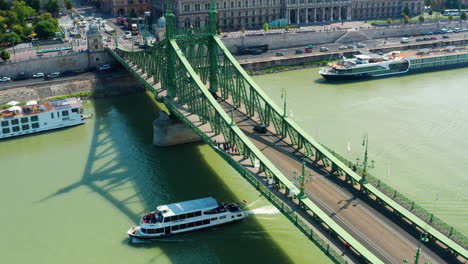 Tourist-boat-sails-under-Liberty-Bridge,-Aerial-pull-back-revealing-Budapest