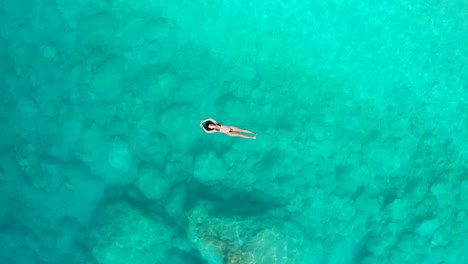 Top-down-view-girl-with-black-bikini-floating-on-her-back-stunning-turquoise-water,-Ascending