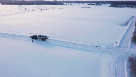 Aerial-view-tractor-trailer-carrying-rolled-bales-across-snowy-winter-rural-agricultural-countryside