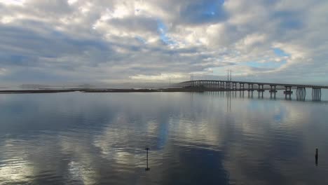 Drone-flight-above-water-with-cloud-reflections-and-bridge-view