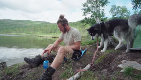 Hombre-Y-Perro-Descansando-Junto-Al-Lago-Preparando-Una-Estufa-De-Camping-Portátil-De-Un-Solo-Quemador-Mientras-Caminan-En-El-Parque-Nacional-De-Ånderdalen-En-La-Isla-De-Senja,-Noruega