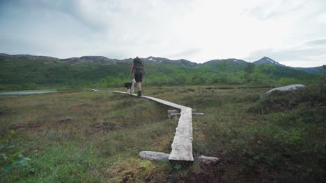 Männlicher-Wanderer-Mit-Einem-Hund,-Der-Auf-Einem-Holzplankenweg-In-Richtung-Eines-Flusses-Im-Anderdalen-nationalpark,-Senja,-Norwegen,-Spazieren-Geht