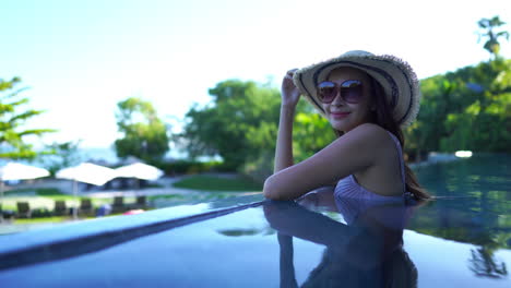 attractive-asian-woman-wearing-a-striped-sunhat-and-large-sunglasses-smiling-as-she-enjoys-a-shady-infinity-pool-with-glassy-water-and-bright-sunlight-on-the-green-trees-in-the-background