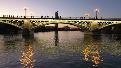 Niedrige-Fliege-Im-Guadalquivir-Fluss,-Der-Unter-Der-Triana-Brücke-Vorbeifährt,-Enthüllt-Den-Schuss-Des-Pelli-Sevilla-Turms-Während-Der-Sonnenuntergangsdämmerung-In-Sevilla,-Spanien
