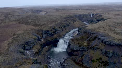 Breathtaking-Aerial-circling-shot-of-waterfall-falling-into-Selá-river-surrounded-by-hills-landscape-during-cloudy-day---North-Iceland-in-autumn-season