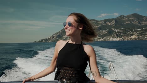 Schönes-Kaukasisches-Mädchen,-Das-Auf-Einer-Fahrenden-Yacht-Mit-Wasserweg-Und-Berghintergrund-In-Italien-Steht