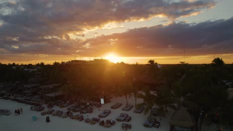 Drone-flying-over-the-houses-surrounded-by-trees,-to-the-Akiin-beach,-with-beautiful-clouds-in-the-background,-at-sunset