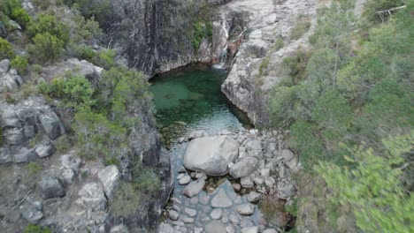 Aerial-pullback-Over-emerald-natural-pool-from-waterfall,-Gerês-National-park,-Portugal
