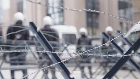 Unrecognizable-riot-police-in-full-riot-gear-backed-by-anti-riot-vans-cordon-off-a-street-during-a-demonstration---defocused-shot