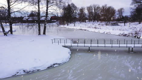 Small-city-park-with-bridge-over-frozen-lake-water-in-winter-season-while-snowing,-aerial-view