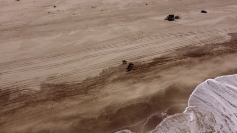 Cinematic-aerial-shot-showing-group-of-people-riding-horses-on-sandy-beach-beside-ocean-during-sunny-day---Mar-de-las-Pampas,Argentina