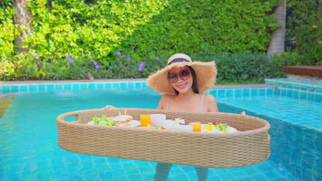 Young-Asian-woman-stands-in-shallow-swimming-pool-with-floating-food-tray